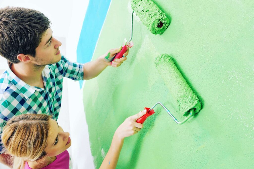 A young couple painting a wall with green paint using paint rollers. The man, wearing a blue and white checkered shirt, and the woman, in a pink top, are working together on the wall. The background shows part of a blue section of the wall.
