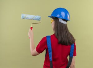 A young female builder wearing a blue safety helmet, yellow safety glasses, and a red shirt with blue suspenders is seen from behind. She is holding a blue paint roller in her right hand, poised as if ready to paint. The background is a plain, light green color.